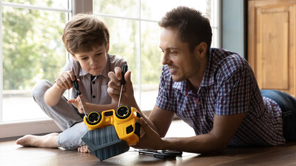 Close up caring father and little son fixing toy car, lying on warm wooden floor at home, smiling young dad and cute adorable boy using screwdriver, having fun, spending leisure time together