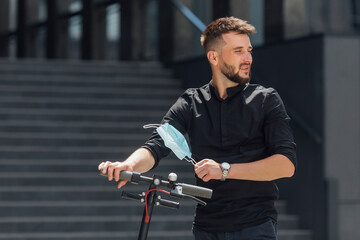 Wall Mural - End of coronavirus epidemic. Happy bearded man taking off medical mask and riding electric scooter in urban background