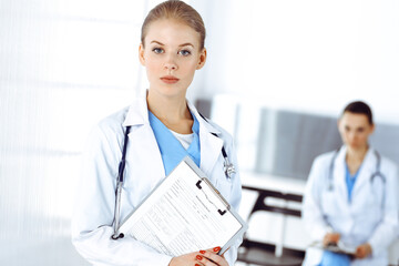 Woman - doctor standing in clinic with colleague at the background. Physician at work. Medicine and health care concept