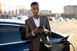 © sofiko14 - Urban, electric vehicle, eco concept. Young black skinned businessman in stylish suit, browsing on cellphone while waiting for his electric car charging