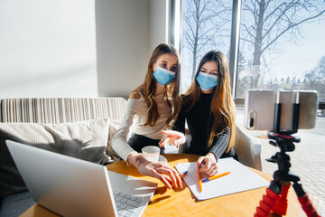 Two young girls sit in a cafe in masks and lead a video blog. Communication to the camera.