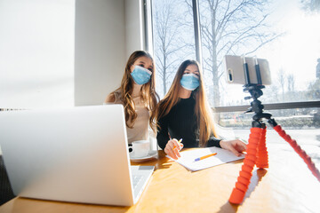 Two young girls sit in a cafe in masks and lead a video blog. Communication to the camera.