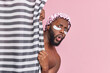 Shocked bearded dark skinned man hides behind shower curtain has amazed face expression applies beauty patches under eyes has daily hygiene procedures isolated over pink background. Cleanliness