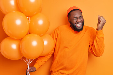 Overjoyed male student celebrates success makes yes gesture being on graduation party holds inflated balloons dressed in casual jumper isolated over orange background. People happiness concept