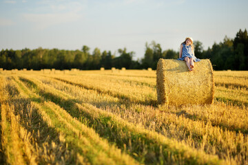 Wall Mural - Adorable young girl having fun in a wheat field on a summer day. Child playing at hay bale field during harvest time. Kid enjoying warm sunset outdoors.