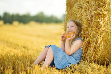 Wall Mural - Adorable young girl eating pretzel in a wheat field on a summer day. Child playing at hay bale field during harvest time. Kid enjoying warm sunset outdoors.