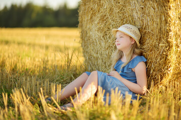 Wall Mural - Adorable young girl having fun in a wheat field on a summer day. Child playing at hay bale field during harvest time. Kid enjoying warm sunset outdoors.