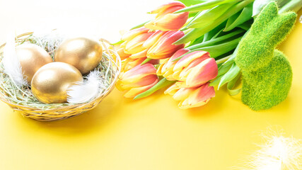 golden eggs in basket with spring tulips, white feathers on pastel yellow background in happy easter