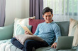 © amnaj - Young Asian man on a leisurely day sitting home on sofa using laptop and tablet on the couch.