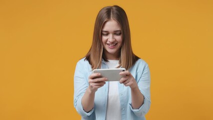 Wall Mural - Joyful young woman in blue shirt isolated on yellow background studio. People lifestyle concept. Using play racing on mobile cell phone hold gadget smartphone for pc video games doing winner gesture