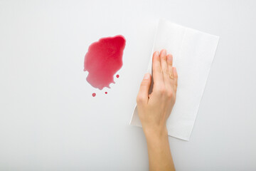 Young adult woman hand cleaning fresh spilled red beverage from light gray table background. Wine stain simple removing with white paper napkin. Closeup. Top down view.