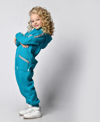 Smiling happy girl in warm fashion jumpsuit trendy sportswear posing for camera on studio copy space. Child beauty model with blond curly hair advertising sportive outerwear. Clothing shop promo