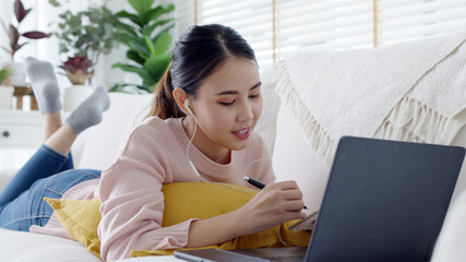 Wall Mural - Cozy relax young attractive asian female employee wear earphone listen to digital online course development program in quarantine social distance at home reskill upskill school class for career path.