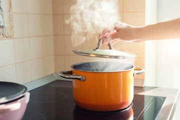 Female hand open lid of enamel steel cooking pan on electric hob with boiling water or soup and scenic vapor steam backlit by warm sunlight at kitchen. Kitchenware utensil and tool at home background