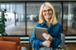 © Kateryna - Joyful blonde-hair business woman in eyeglasses is looking at the camera, holding laptop in arms and smiling. Beautiful middle aged lady in stylish wear stands in modern office