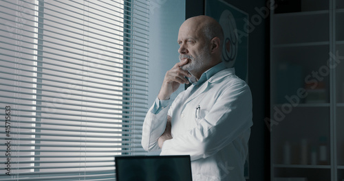 Pensive doctor standing next to a window © stokkete