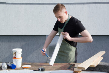 A young male designer paints a wooden Board white with a brush. The young man is working near his house. 