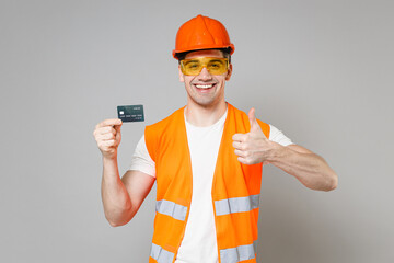 Poster - Young employee man in orange vest protective helmet hold in hand credit bank card show thumb up gesture isolated on grey background studio. Instruments for renovation apartment Repair home concept.