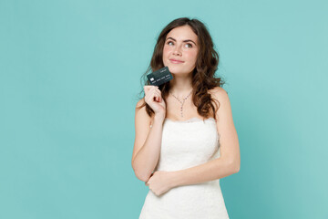 Pensive bride young woman 20s in beautiful white wedding dress hold credit bank card looking aside up isolated on blue turquoise color background studio portrait. Ceremony celebration party concept.