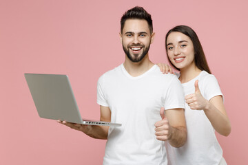 Young couple two friends man woman user in white basic blank print design t-shirts hold using work on laptop pc computer freelance remote jobs isolated on pastel pink color background studio portrait.