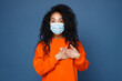 canvas print picture - Pretty young african american woman in casual orange sweatshirt sterile face mask to safe from coronavirus virus covid-19 holding hands on heart isolated on blue color background studio portrait.