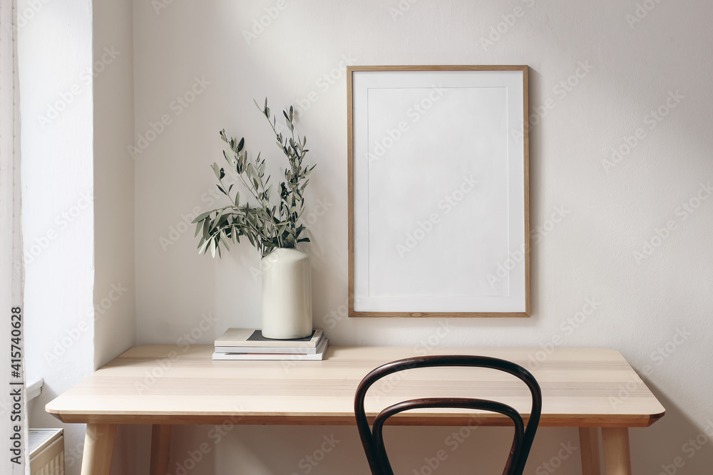 Home office concept. Old books, empty vertical wooden picture frame mockup hanging on white wall. Wooden desk, table. Vase with olive branches. Elegant working space. Scandinavian interior design. - obrazy, fototapety, plakaty 