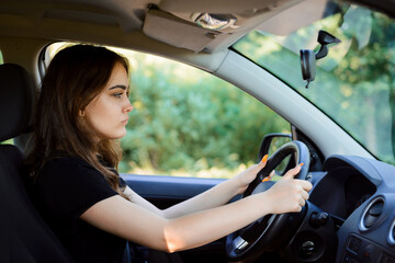 Wall Mural - Young sad girl driving car on a summer day. Female driver with a bad mood inside the automobile.