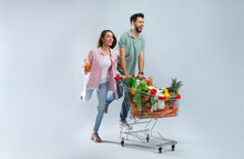 Young Couple With Shopping Cart Full Of Groceries On Grey Background