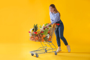 Poster - Young woman with shopping cart full of groceries on yellow background