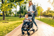 Young Mother Walks With A Little Toddler In A Stroller In The Summer On Nature