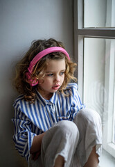 Wall Mural - Sad small girl with headphones indoors at home, sitting on window sill.