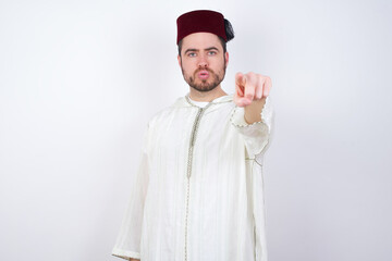 Wall Mural - Cheerful young handsome Caucasian man wearing Arab djellaba and Fez hat over white wall indicates happily at you, chooses to compete, has positive expression, makes choice.