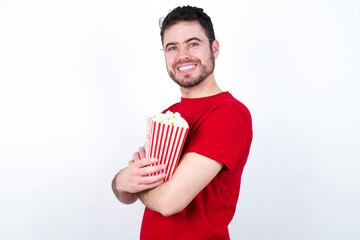 Wall Mural - Image of cheerful young handsome man in red T-shirt against white background eating popcorn with arms crossed. Looking and smiling at the camera. Confidence concept.