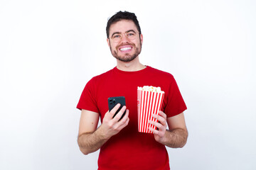 Wall Mural - Smiling young handsome man in red T-shirt against white background eating popcorn friendly and happily holding mobile phone taking selfie in mirror.