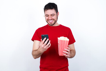 Wall Mural - Smiling Young handsome man in red T-shirt against white background eating p using cell phone, messaging, being happy to text with friends, looking at smartphone. Modern technologies and communication.