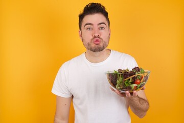 Wall Mural - young handsome Caucasian man holding a salad bowl against yellow wall, keeps lips as going to kiss someone, has glad expression, grimace face. Standing indoors. Beauty concept.