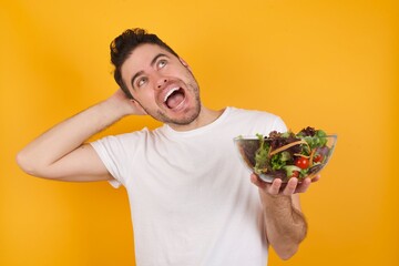 Wall Mural - young handsome Caucasian man holding a salad bowl against yellow wall relaxing and stretching, arms and hands behind head and neck smiling happy