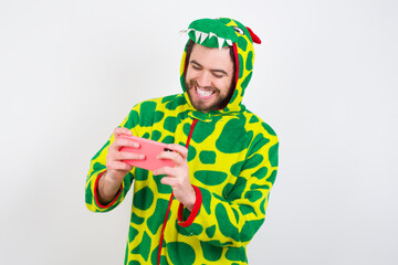 Sticker - young caucasian man wearing a pajama standing against white background holding in hands cell playing video games or chatting