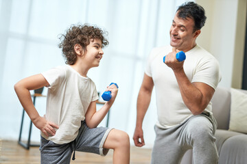 Shaping bodies. Sportive latin middle aged father teaching his son exercising with dumbbells while spending time together at home