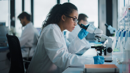 medical science laboratory: beautiful black scientist looking under microscope does analysis of test