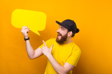 Wall Mural - Charming young bearded man pointing at yellow speech bubble.