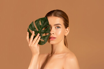 Wall Mural - Portrait of pretty young woman with smooth skin and nude makeup closing her eye with monstera leaf on brown background