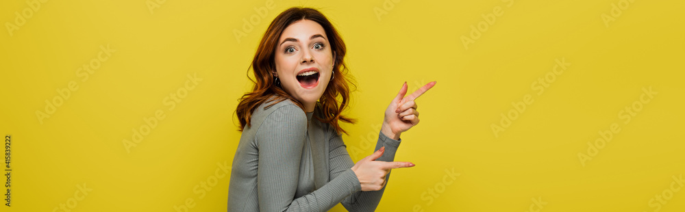 excited woman looking at camera while pointing with fingers isolated on yellow, banner - obrazy, fototapety, plakaty 