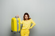 © LIGHTFIELD STUDIOS - excited woman holding yellow suitcase while standing with hand on hip on grey