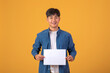 © amnaj - Young Asian man on orange background Living concept. Mock up copy space. Hold a blank paper. Looking at camera.