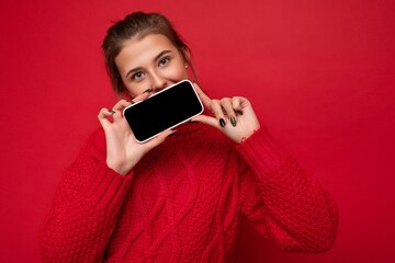 Photo of beautiful cute young woman wearing warm red sweater isolated over red background wall holding smartphone and showing phone with empty display for mockup looking at camera