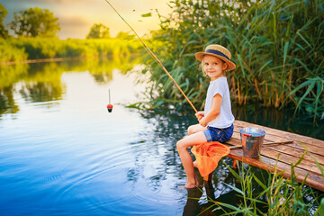 Wall Mural - Little boy in straw hat sitting on the edge of a wooden dock and fishing in lake at sunset.