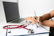 © Chinnabuth - doctor working on laptop taking notes at workplace. Female physician writing in notebook using computer sitting at desk.