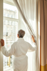 Wall Mural - Back view of mature woman in bathrobe pulls the curtain and looks out while standing in hotel room