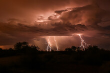 Lightning Strike During Summer Thunderstorm, In Arizona's Sonoran Desert. Three Bolts Extend Down From The Clouds, Which Are Backlit By The Discharge. Silhouettes Of Brush In The Foreground.
